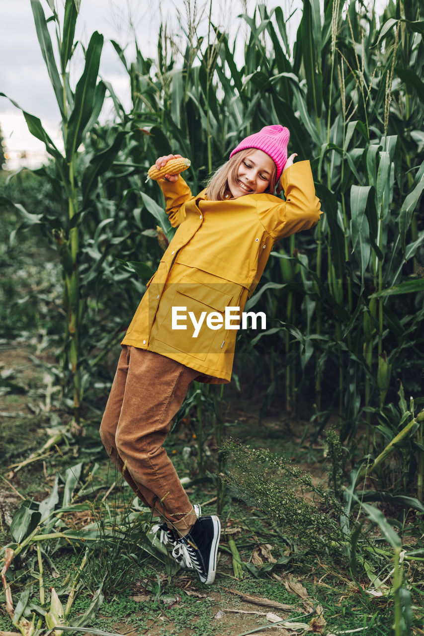 Stylish teenage girl in a yellow raincoat and a hot pink cap is dancing and laughing on a cornfield