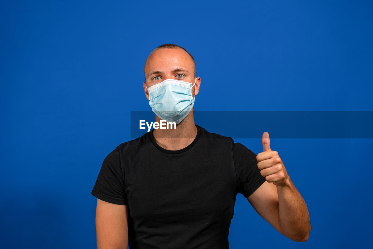 blue, adult, one person, men, studio shot, portrait, healthcare and medicine, colored background, arm, front view, human face, indoors, blue background, person, looking at camera, waist up, copy space, clothing, protective mask - workwear, standing, surgical mask, gesturing, shaved head