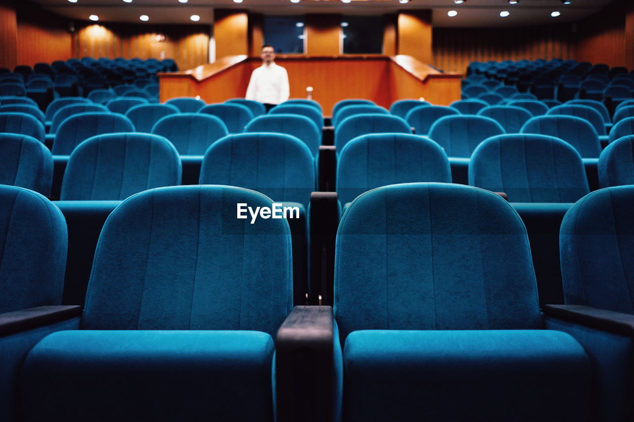 Defocused mid adult man standing amidst blue empty chairs in auditorium