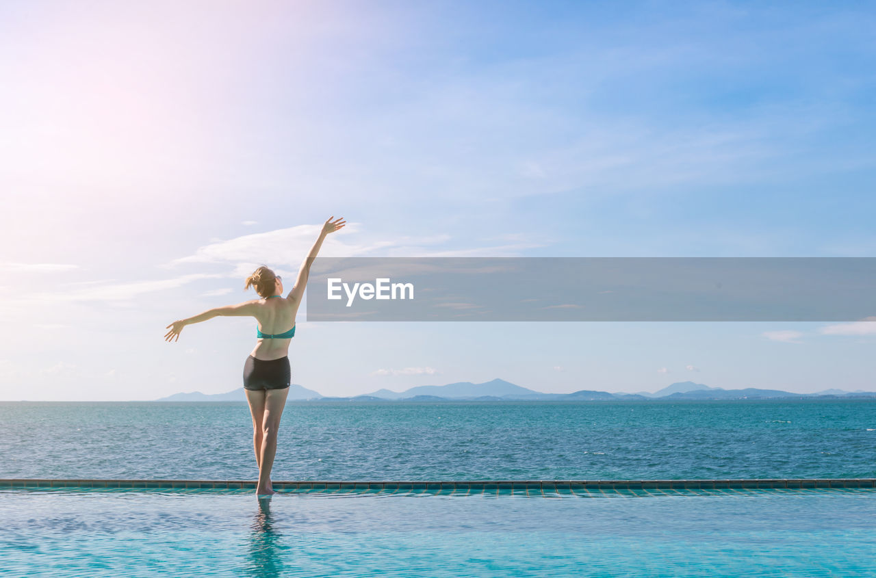 Rear view of woman with arms raised standing at infinity pool by sea