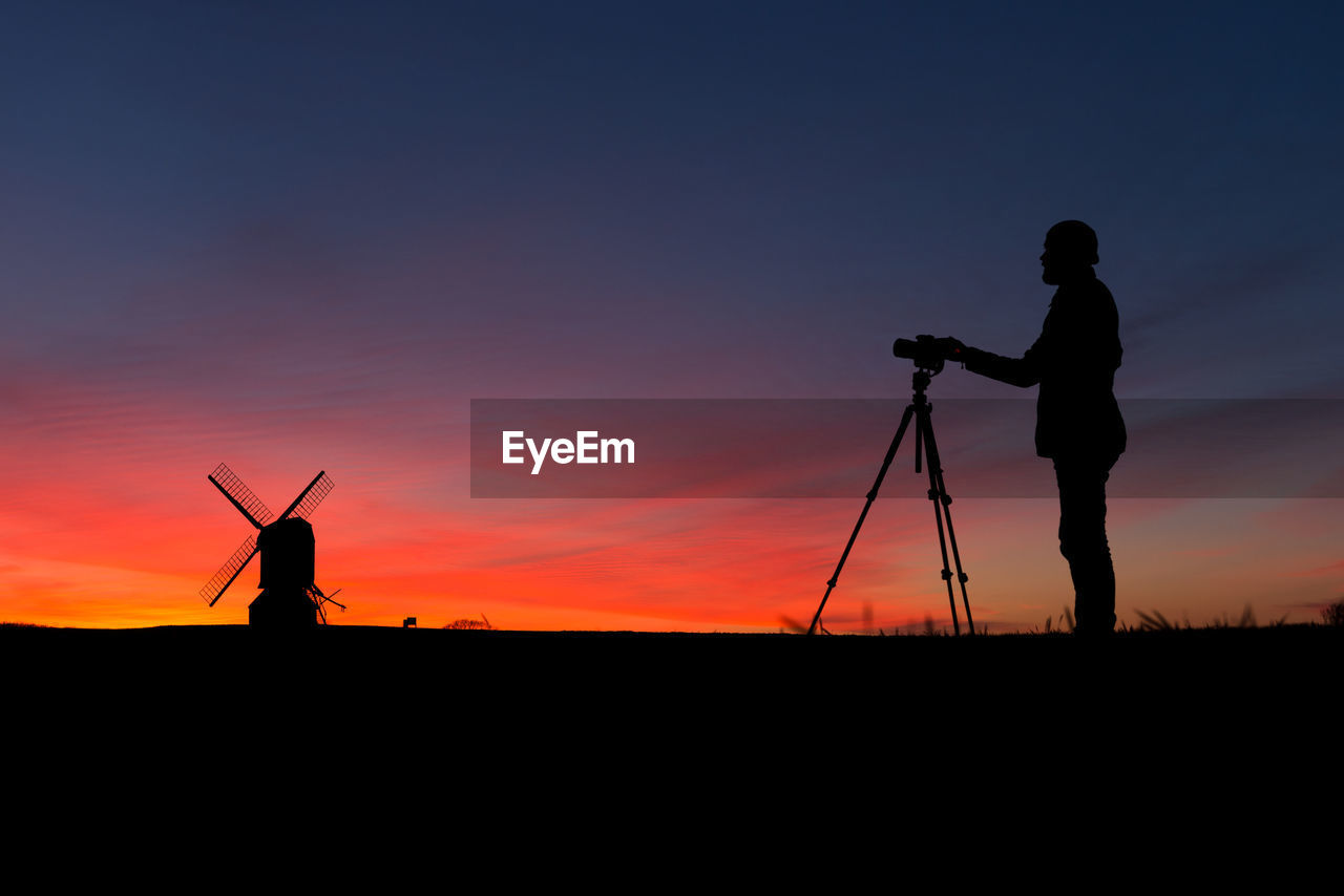 Silhouette photographer with tripod by windmill against sky at dusk