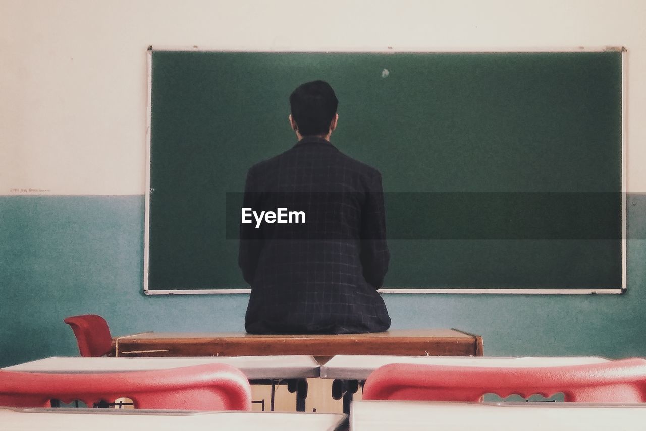 Rear view of man sitting on table looking at blackboard