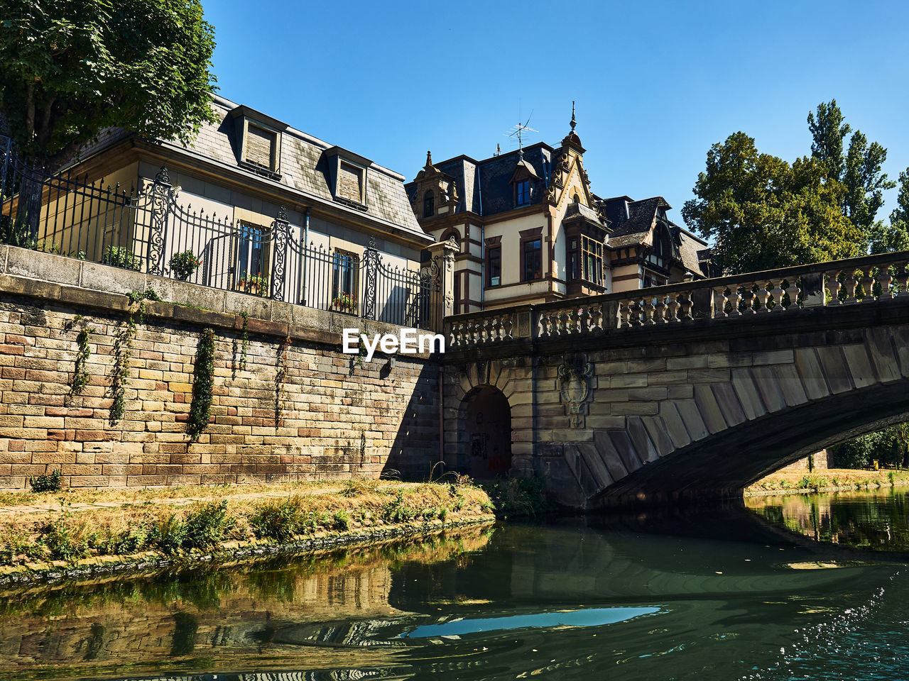 architecture, built structure, water, river, bridge, waterway, building exterior, nature, travel destinations, history, sky, the past, travel, city, landmark, tree, tourism, reflection, clear sky, building, plant, moat, château, transportation, no people, town, blue, waterfront, sunny, outdoors, arch, day, old, estate, arch bridge, medieval