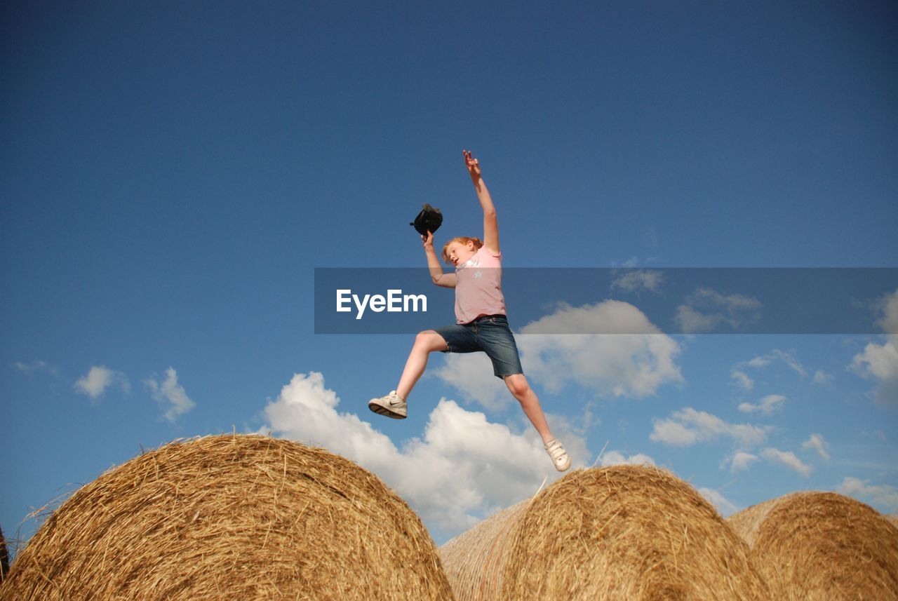 Low angle view of young woman jumping on hay bales against sky