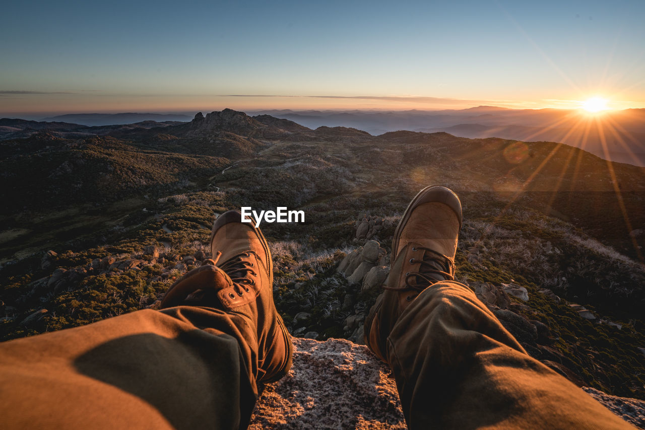 Low section of man sitting on cliff against sky during sunset