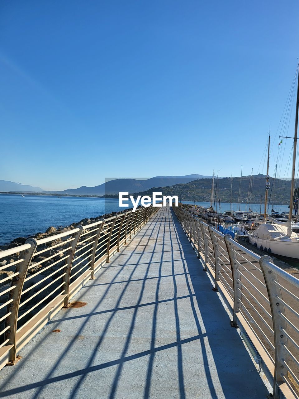 transportation, sky, water, sea, nature, dock, walkway, blue, clear sky, no people, mode of transportation, nautical vessel, travel, ship, the way forward, day, sunlight, coast, scenics - nature, horizon, beauty in nature, sunny, vehicle, marina, travel destinations, sailboat, architecture, outdoors, boardwalk, tranquility, railing, pier, tranquil scene, ocean, copy space, diminishing perspective, bridge, mountain, land, vacation, sailing, tourism, beach, mast, vanishing point, harbor