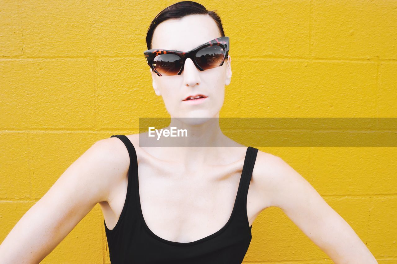 Young woman wearing sunglasses standing against yellow wall