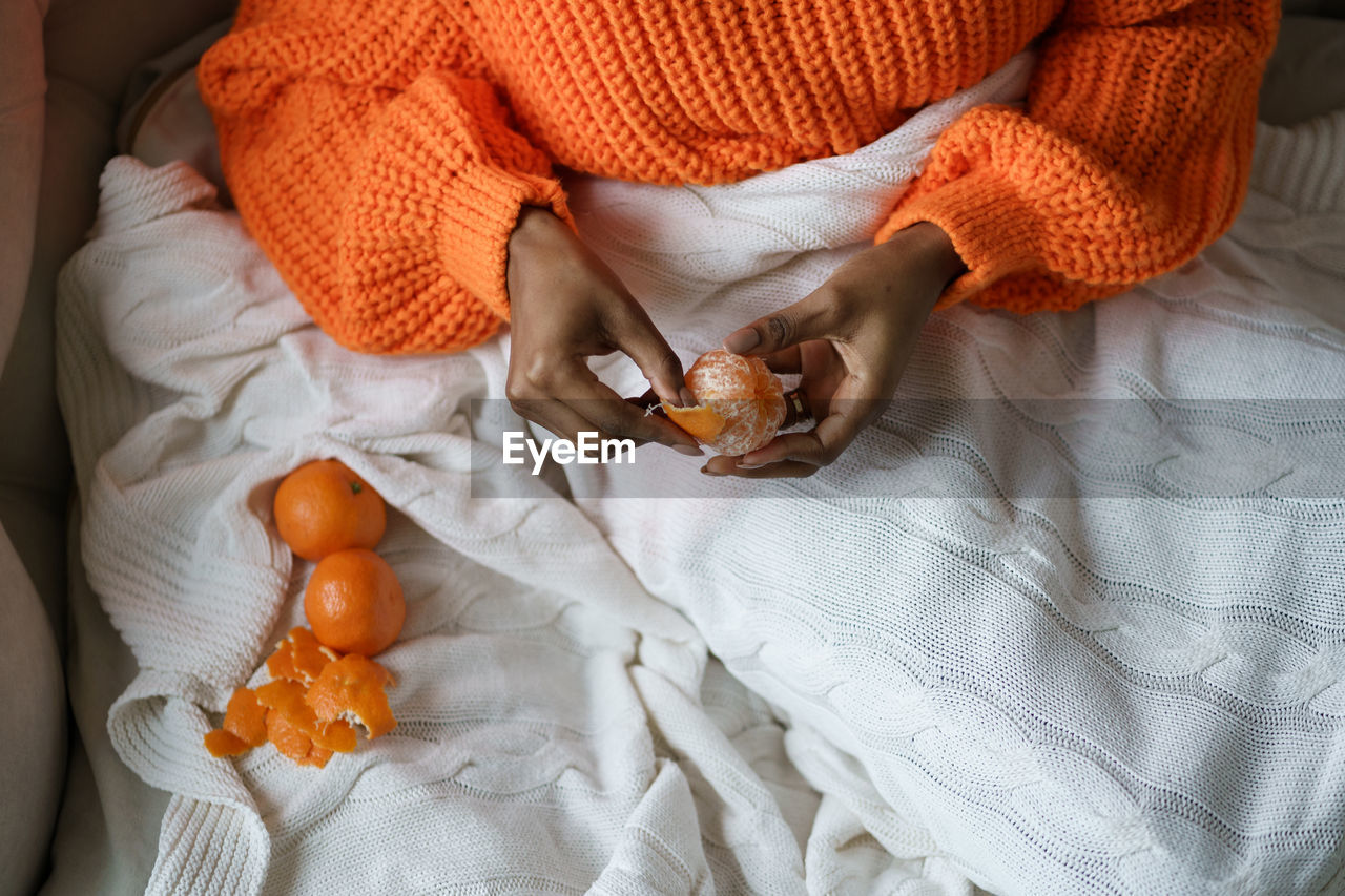 Afro woman hands peeling ripe sweet tangerine, wear orange sweater, lying in bed under the blanket