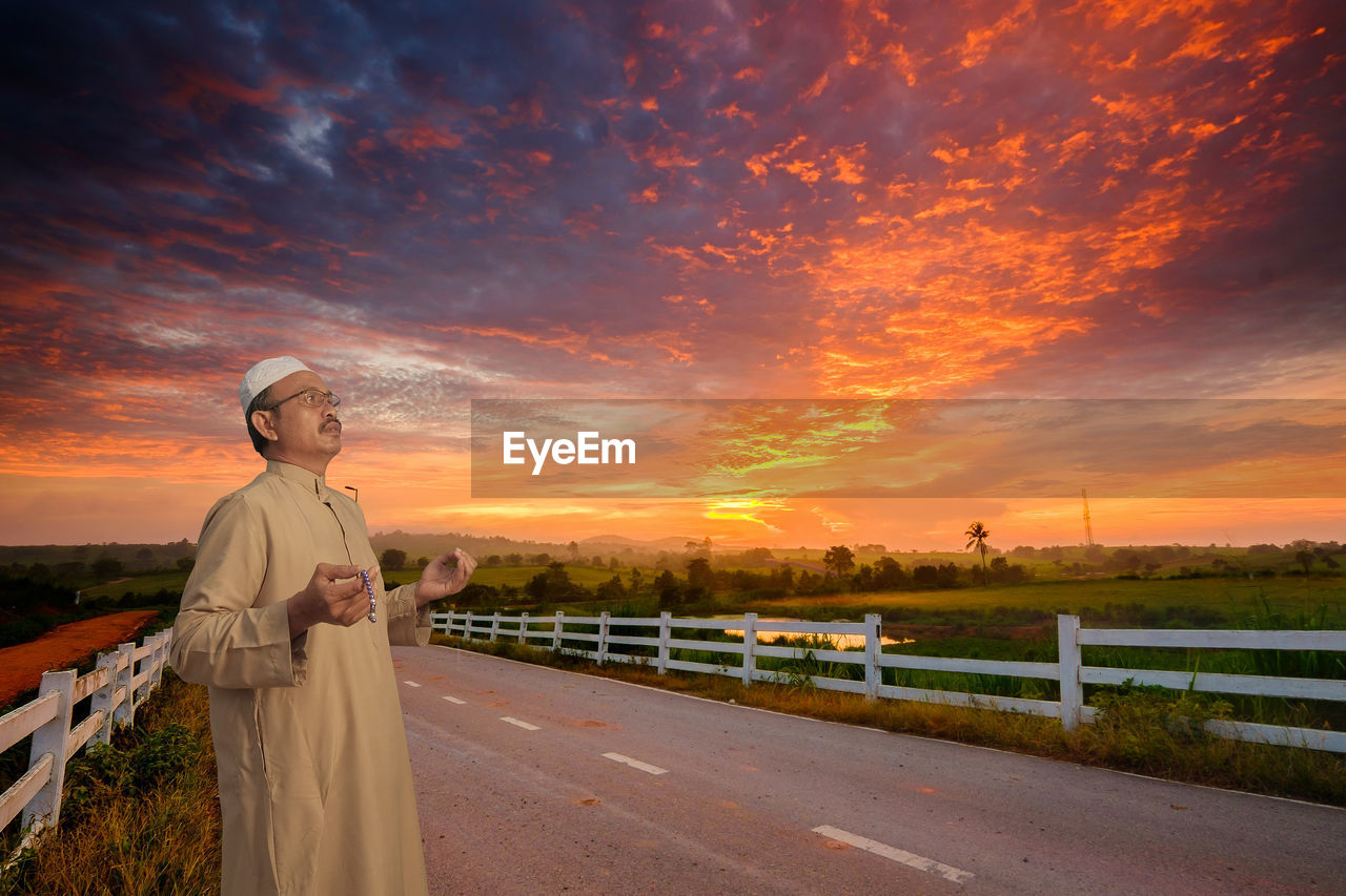 Mature man wearing taqiyah praying while standing on road against dramatic sky during sunset