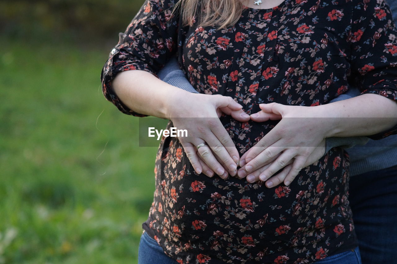 Pregnant couple making heart shape with hand over abdomen while standing on grass