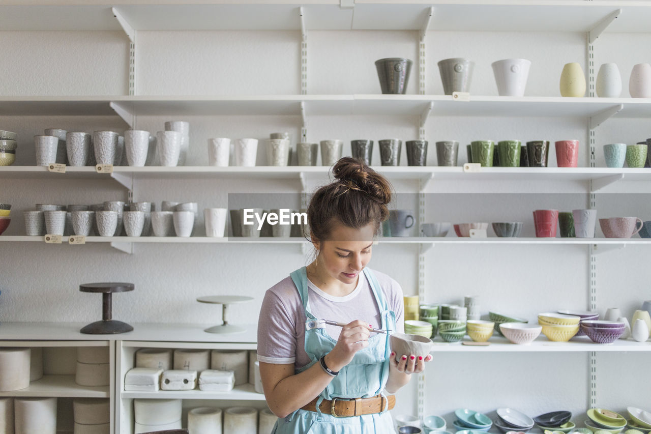 Young female potter using hand tool on vase while standing against shelves at store