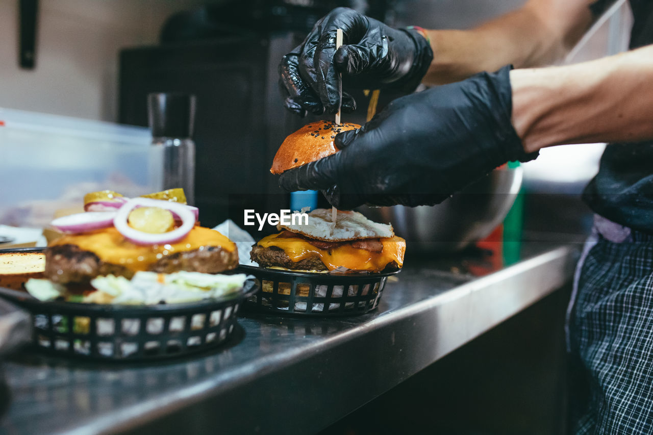 Side view of crop anonymous cook in black gloves arranging stick into freshly made hamburger while preparing order at kitchen counter