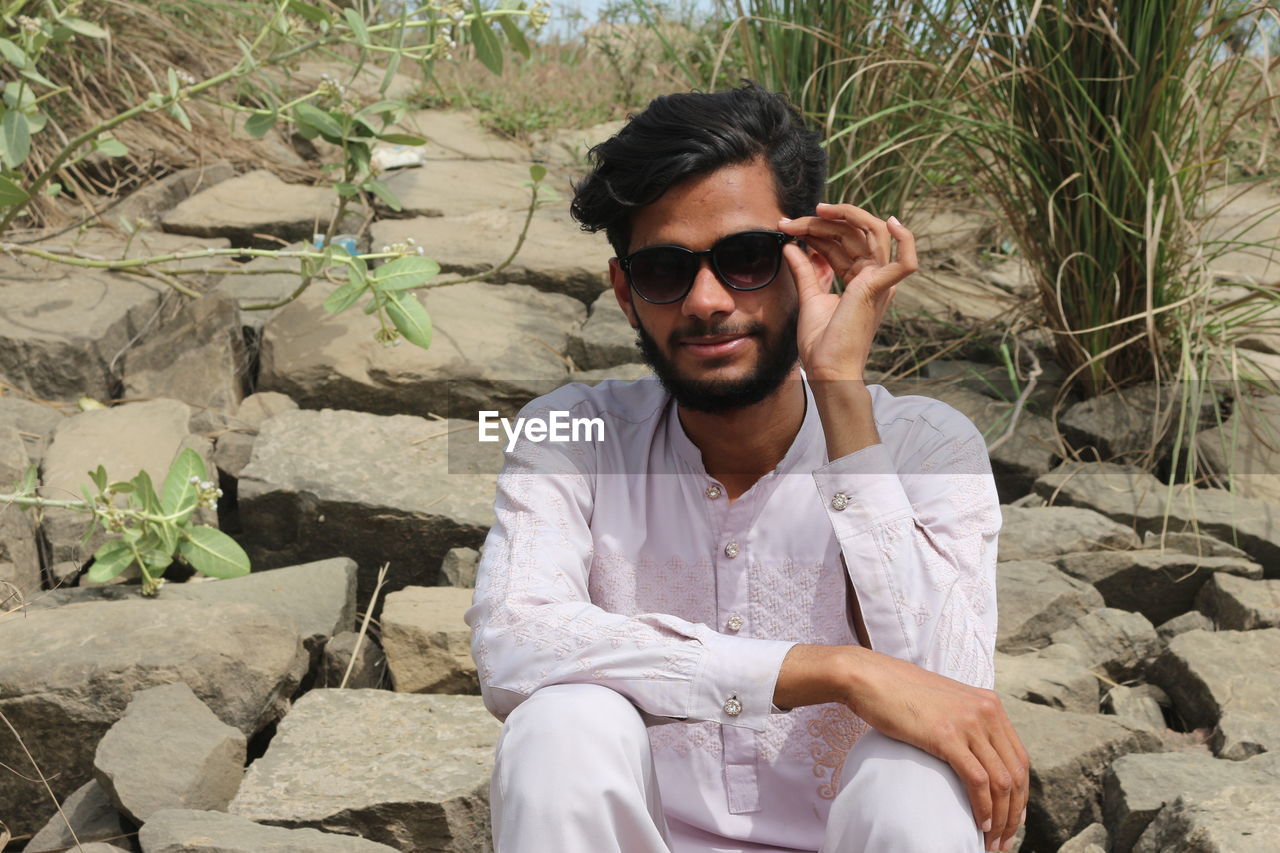 one person, sunglasses, sitting, young adult, glasses, front view, fashion, leisure activity, nature, adult, casual clothing, men, lifestyles, rock, beard, day, land, relaxation, portrait, facial hair, three quarter length, person, outdoors, plant, sunlight, smiling, looking at camera, clothing, black hair, spring, vacation, beach, trip