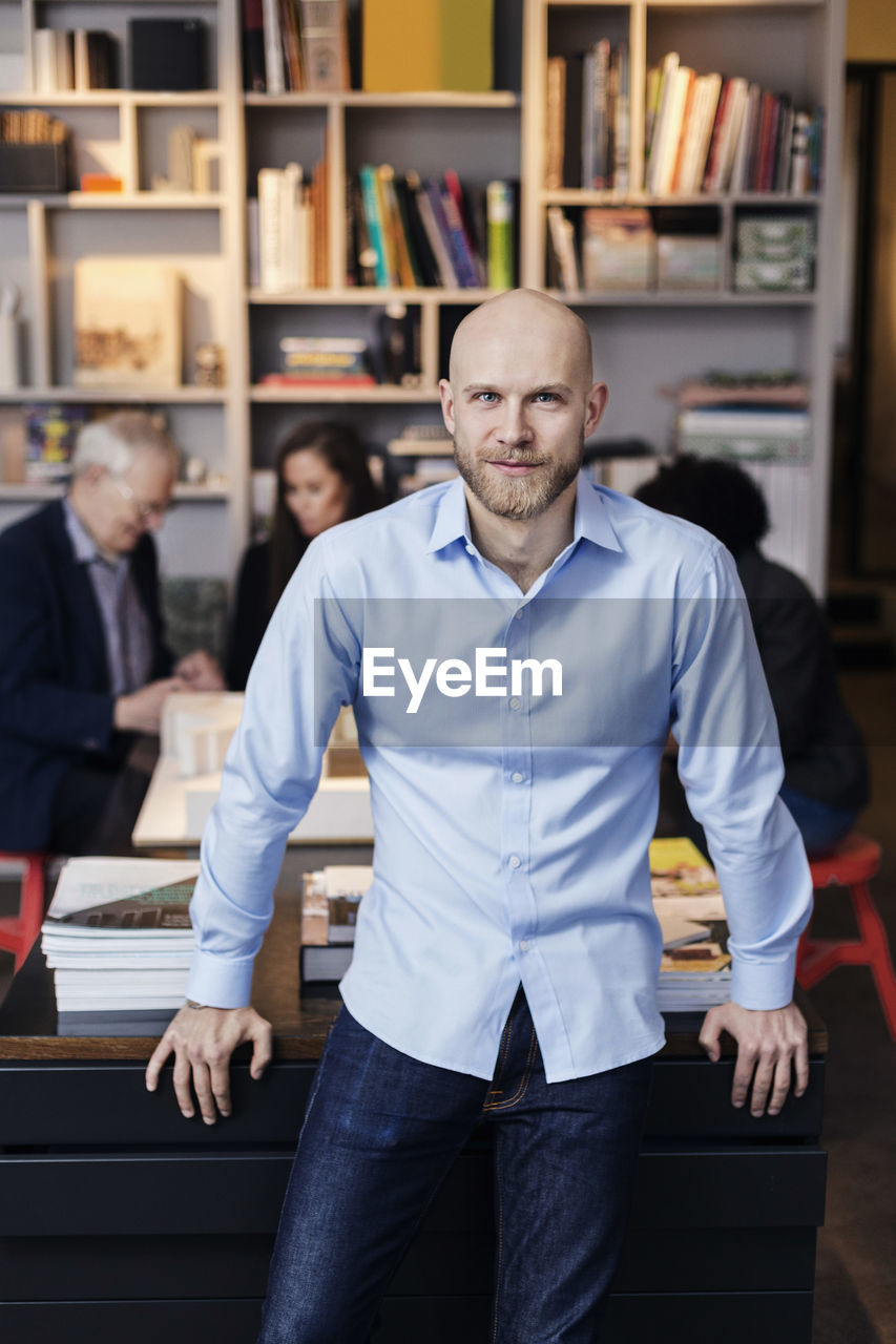 Portrait of confident businessman leaning on desk while colleagues working in background