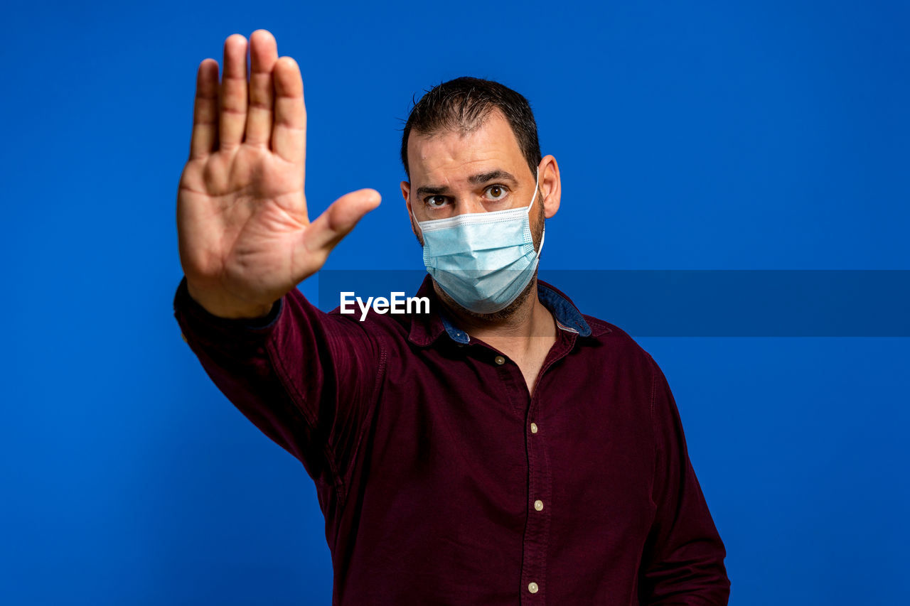 one person, blue, men, adult, studio shot, hand, person, finger, gesturing, portrait, colored background, human face, arm, sign language, looking at camera, indoors, healthcare and medicine, blue background, clothing, glasses, human head, front view, waist up, communication, protection, emotion, security, sign, young adult