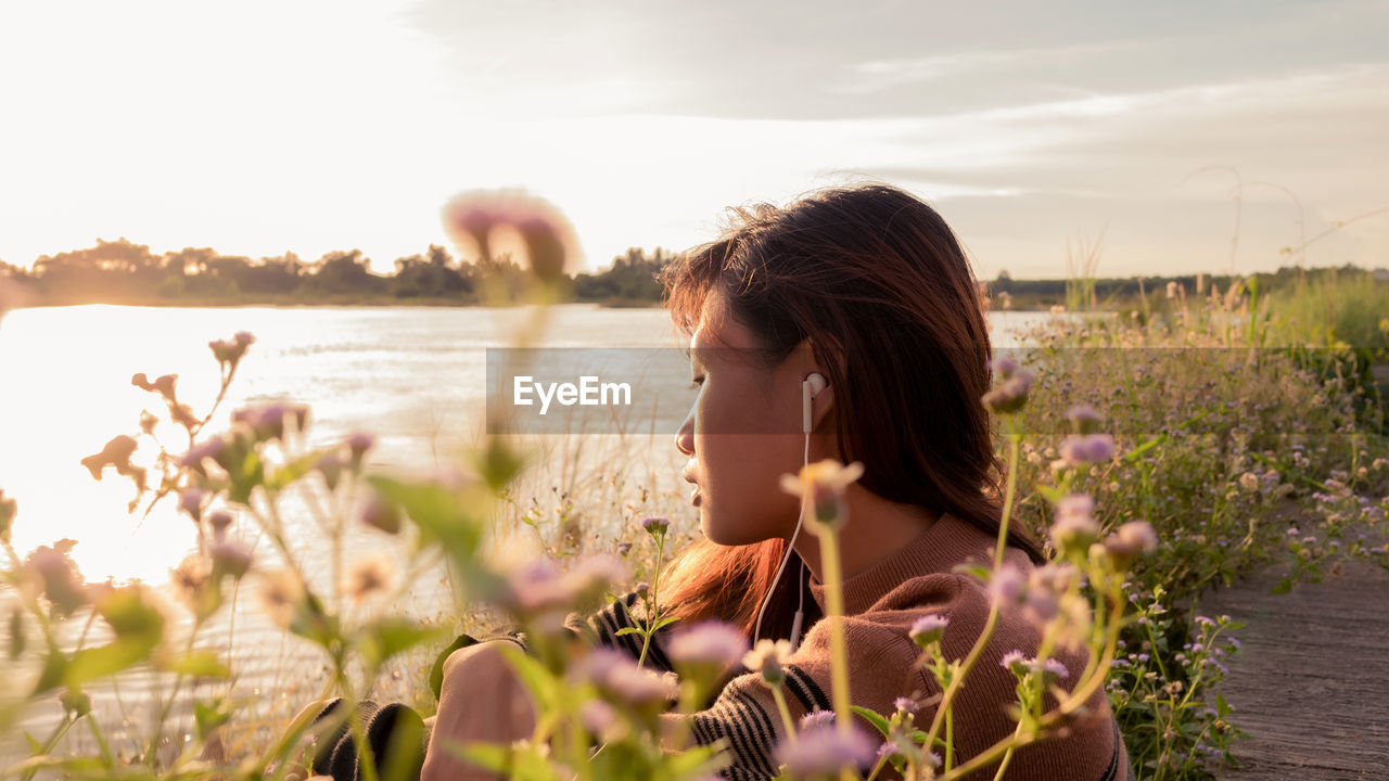 PORTRAIT OF WOMAN ON FLOWERING PLANTS AGAINST SKY DURING SUNSET