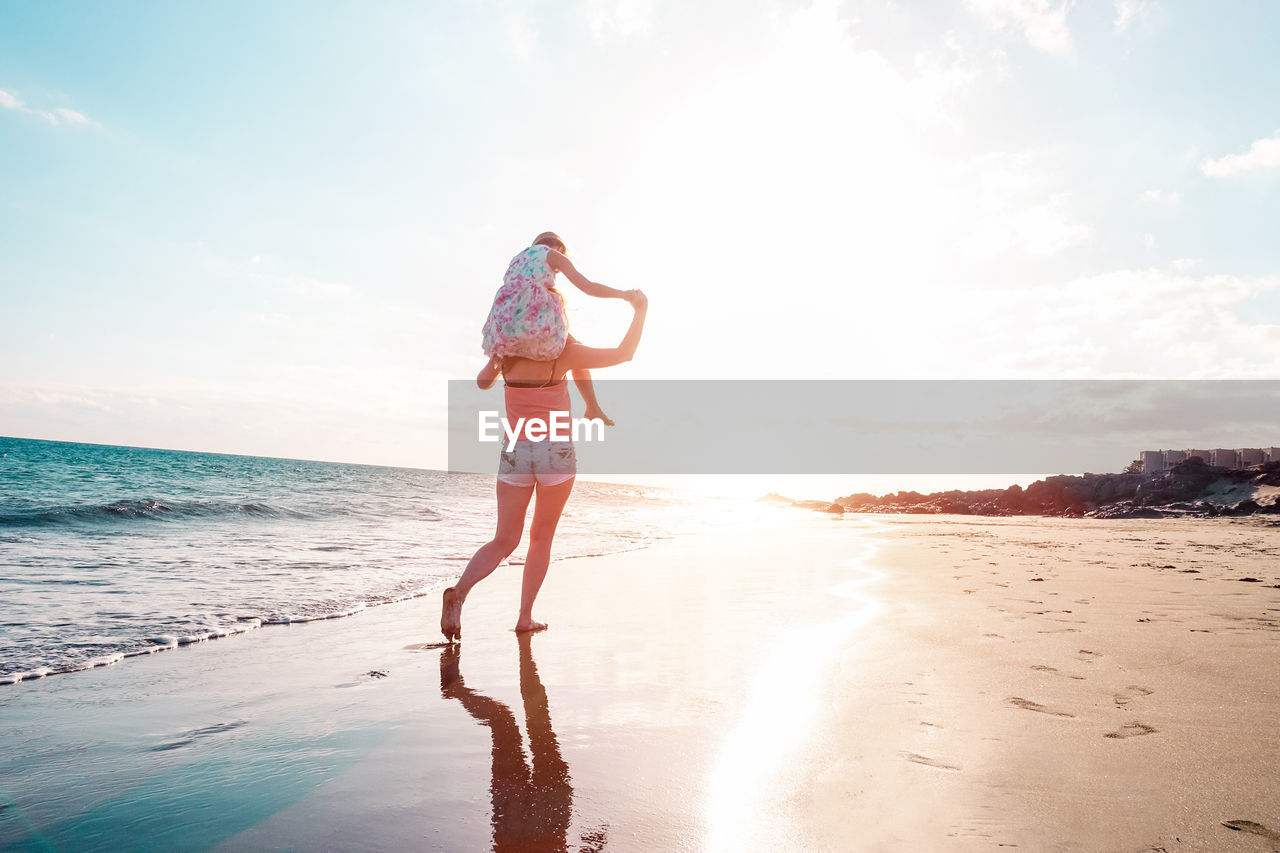Rear view of mother carrying daughter on shoulders at beach against sky during sunset
