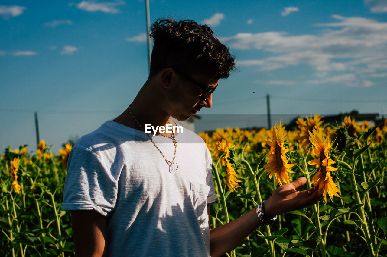 Young man touching sunflower on field against sky during sunny day