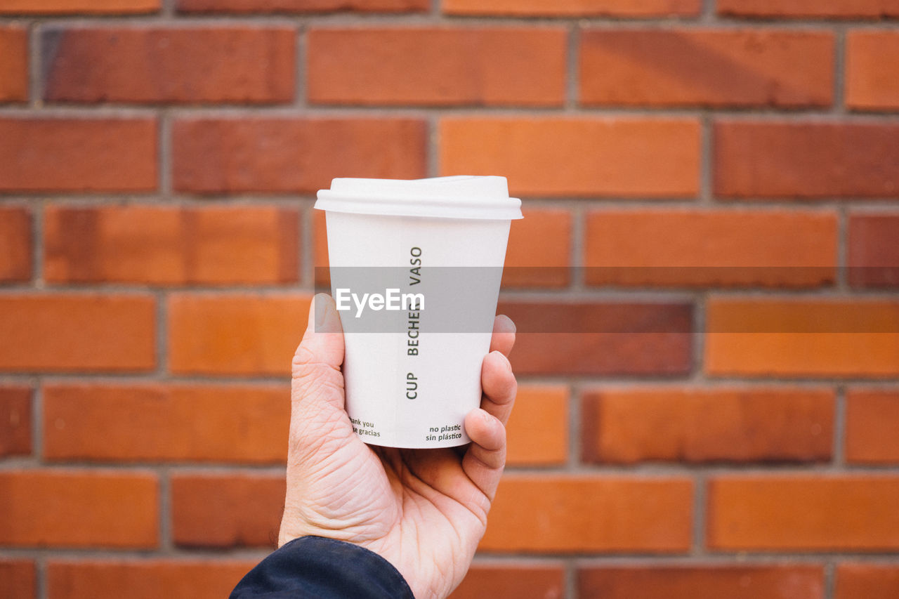 Cropped hand holding coffee cup in front of brick wall