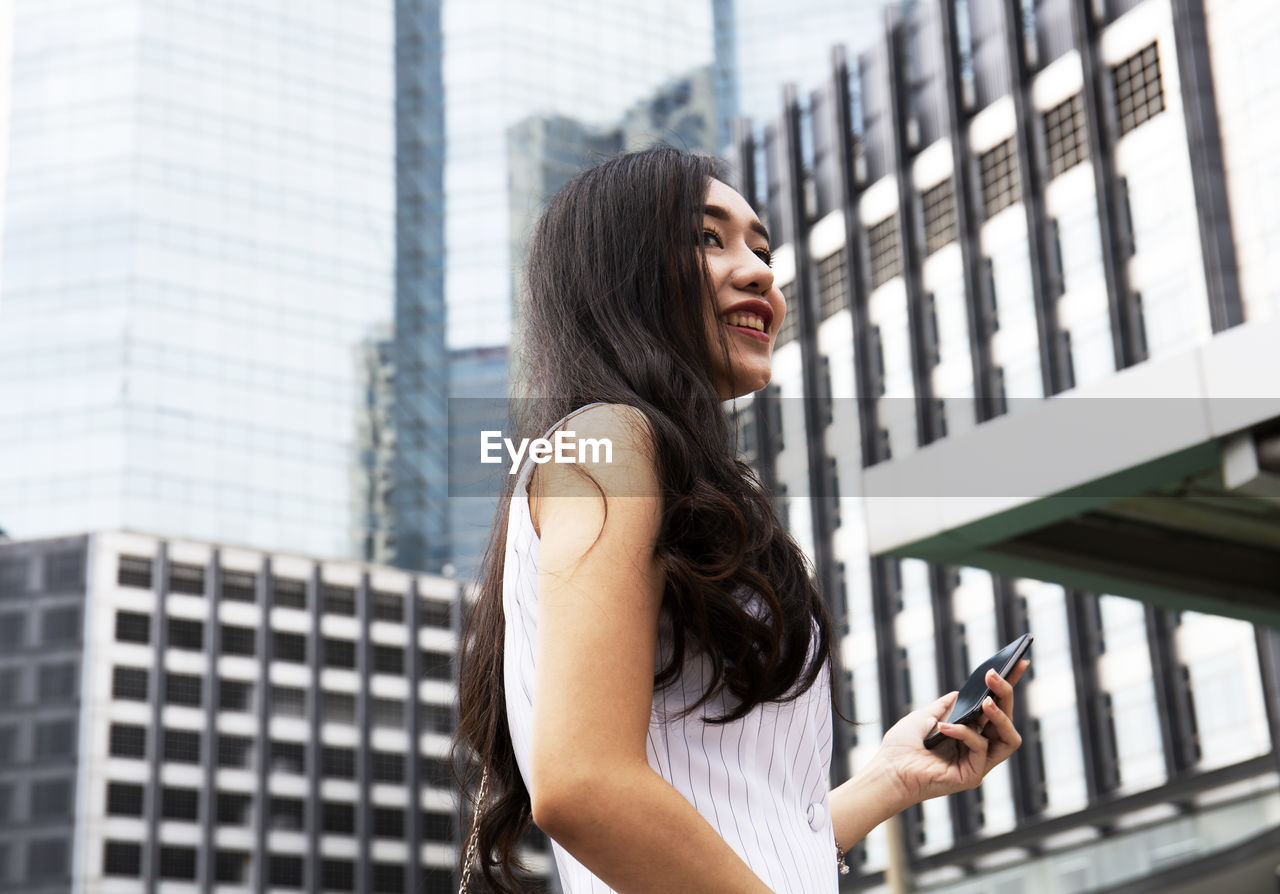 Smiling young woman holding mobile phone while standing in city