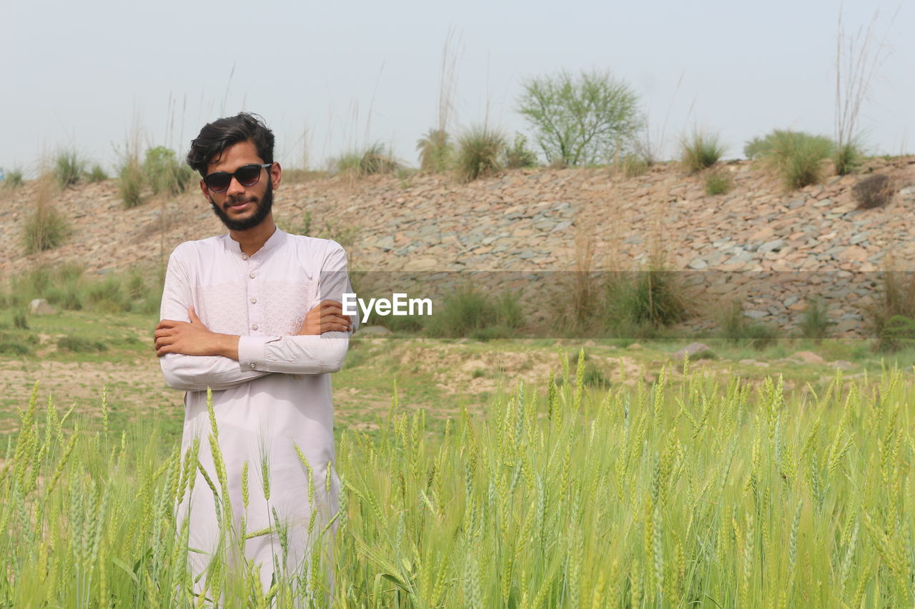one person, plant, adult, field, nature, agriculture, natural environment, men, grass, glasses, land, sunglasses, prairie, landscape, fashion, front view, rural area, portrait, grassland, smiling, young adult, rural scene, three quarter length, crop, day, casual clothing, looking at camera, environment, standing, occupation, growth, sky, arms crossed, outdoors, meadow, leisure activity, happiness, facial hair, lifestyles, beard, tranquility, clothing, emotion, beauty in nature, copy space, cereal plant