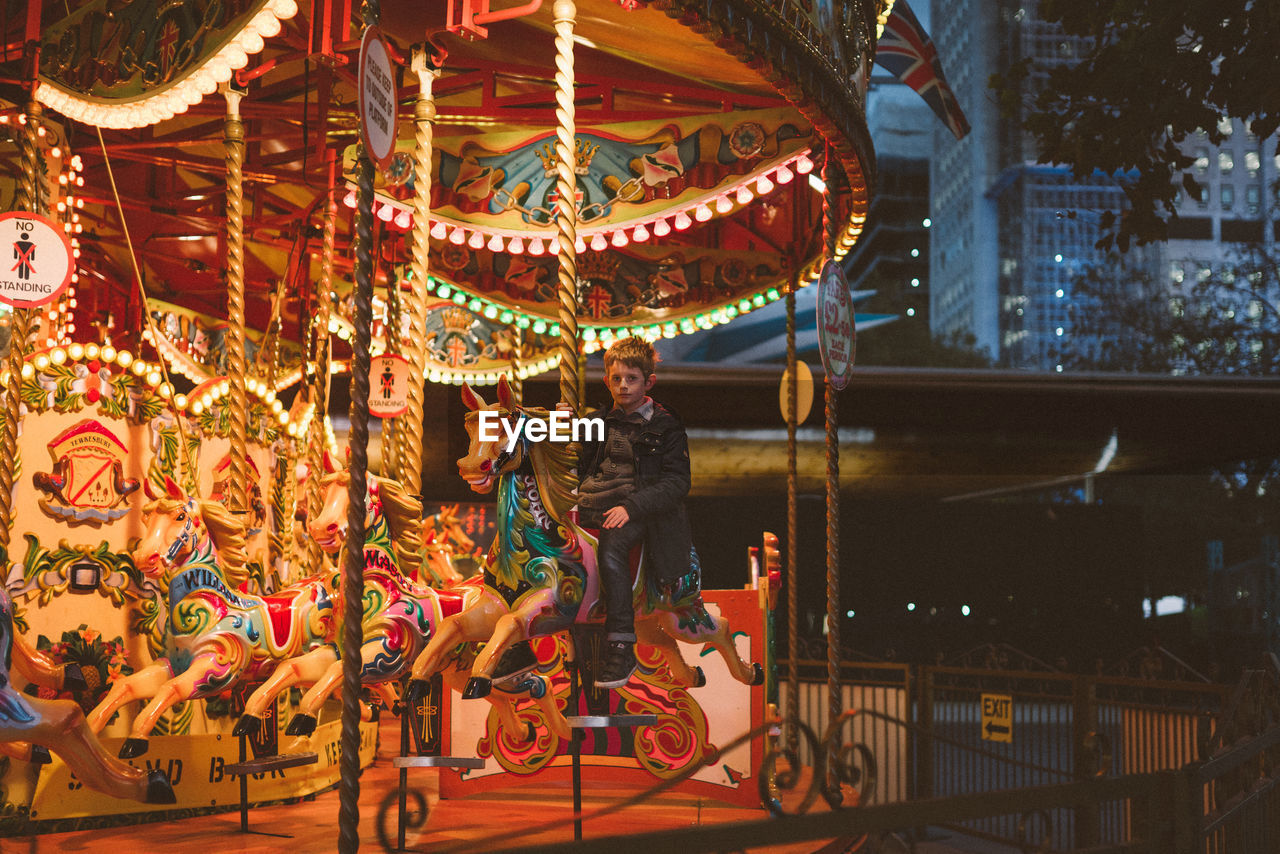 Boy sitting on illuminated carousel in amusement park