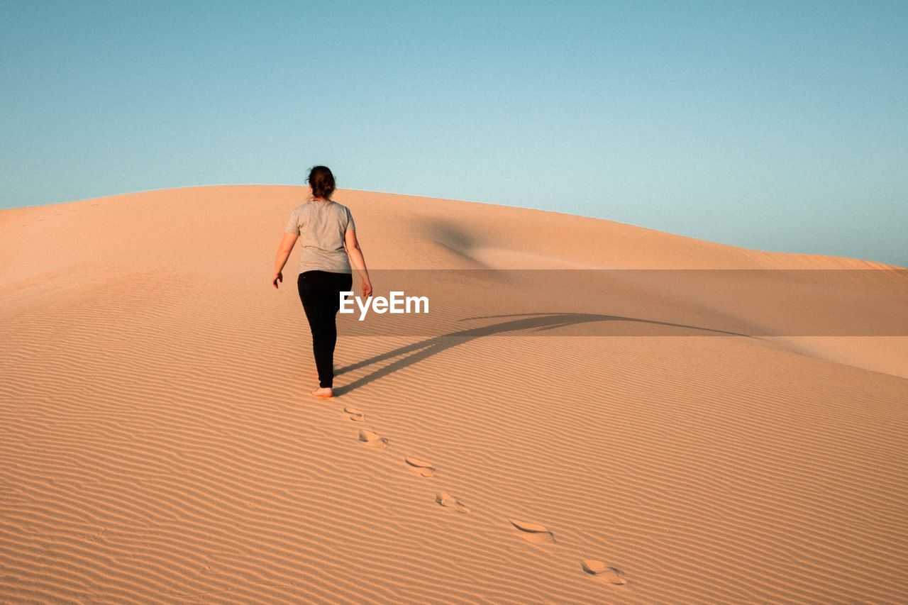 Rear view of woman walking on sand dune at desert against clear sky