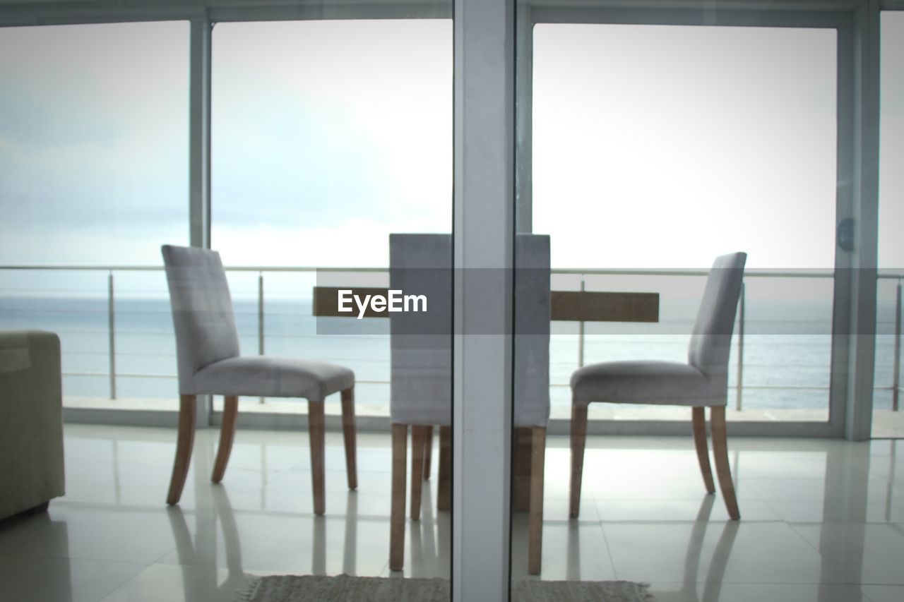 Empty chairs on balcony by sea seen through glass door