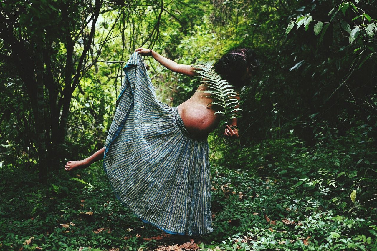 Shirtless pregnant woman holding leaves while standing in forest