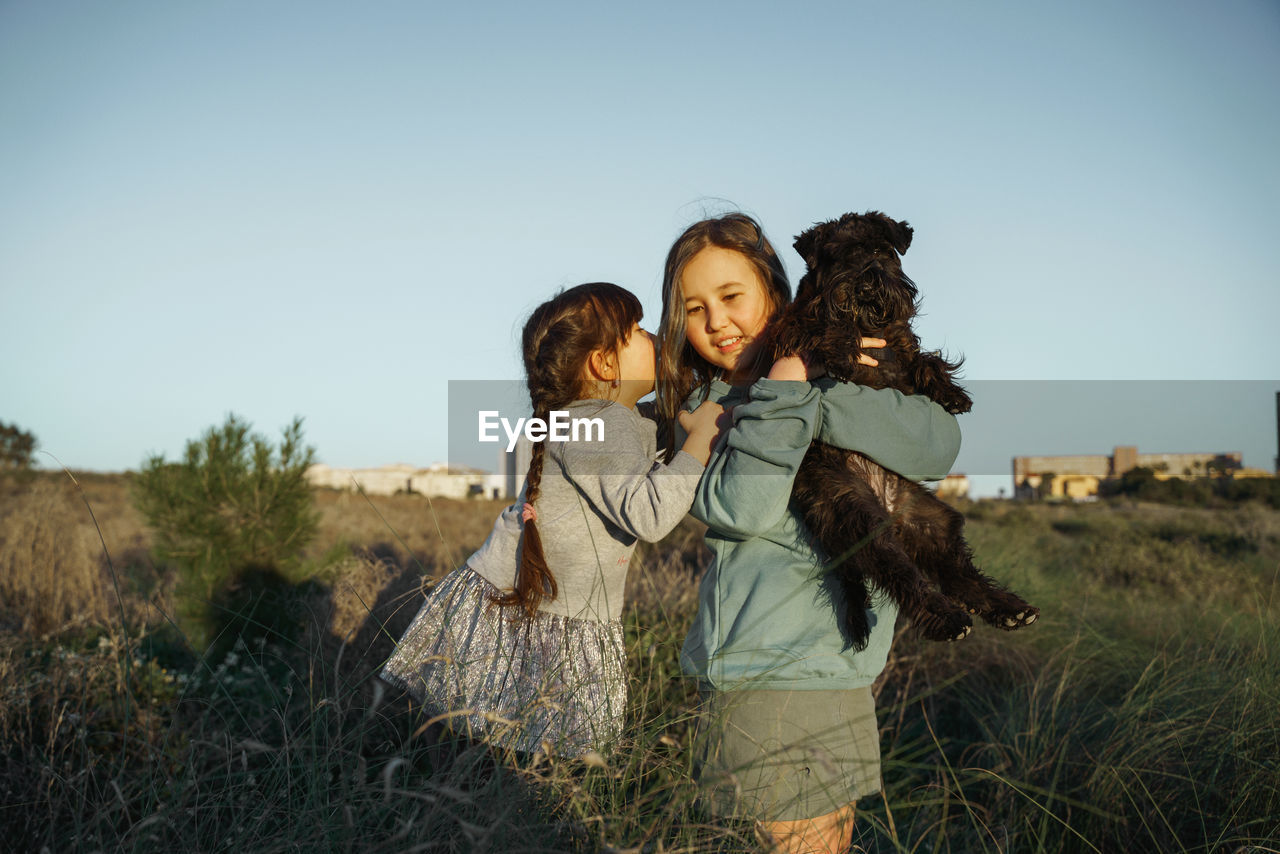 Two girl standing on field with dog against clear sky