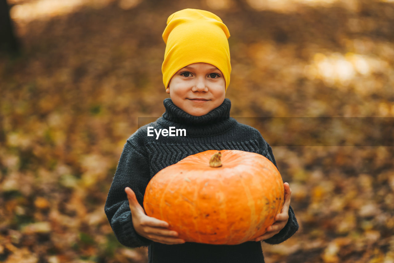 Happy funny little boy teen child in a hat holding a big pumpkin is getting ready for halloween