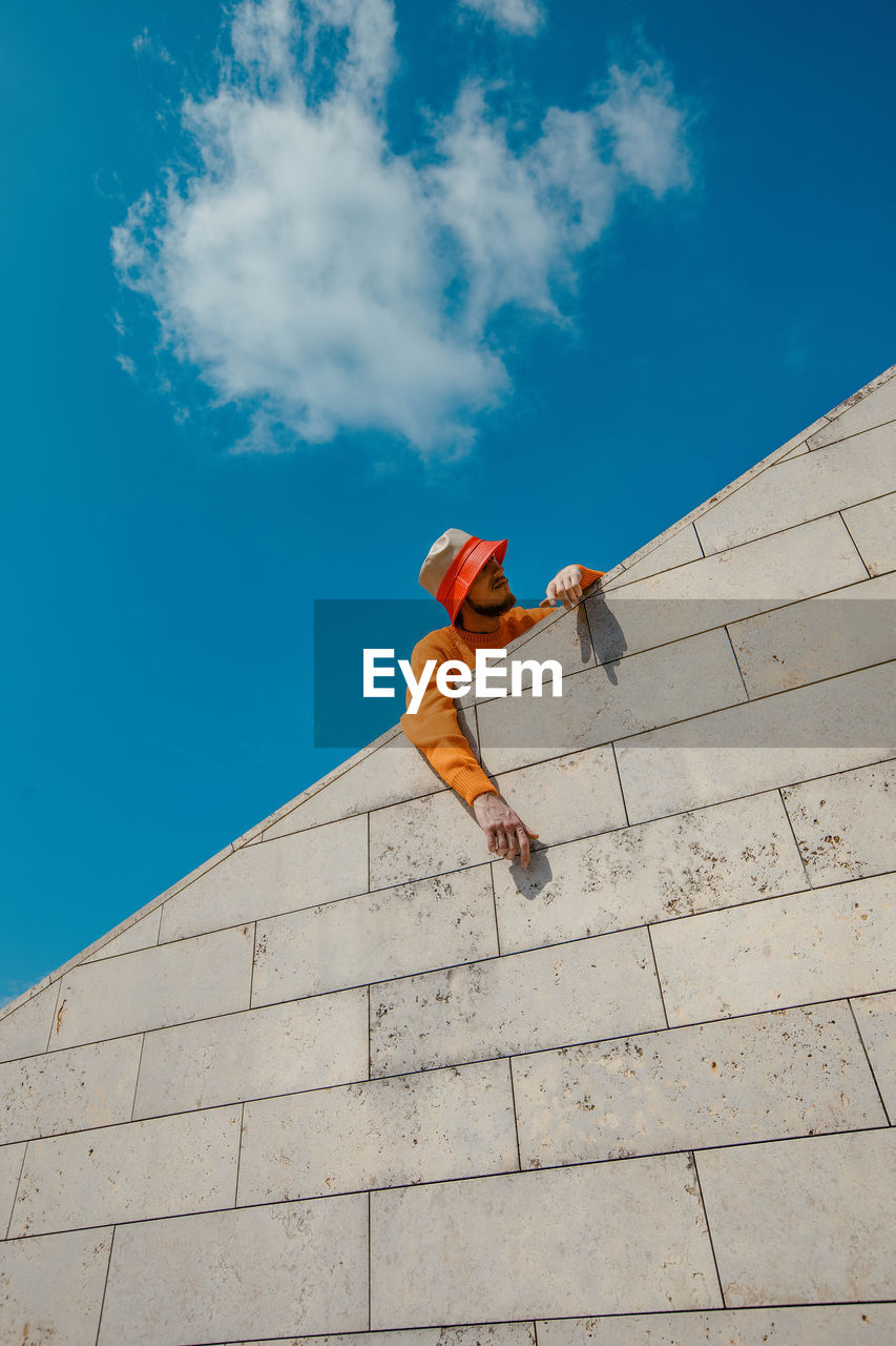 Low angle view of man standing by retaining wall against blue sky