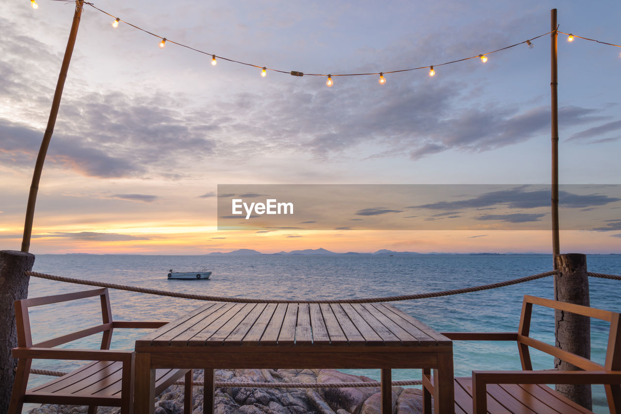 Empty wooden table and seats on pier over sea against sky during sunset