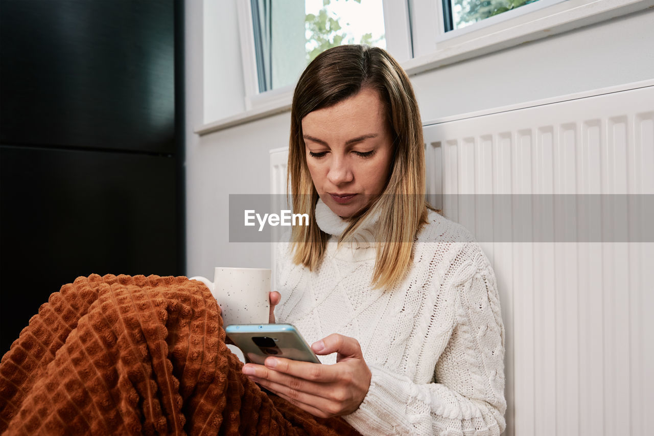 Worried woman sit near heating radiator under blanket and using smartphone