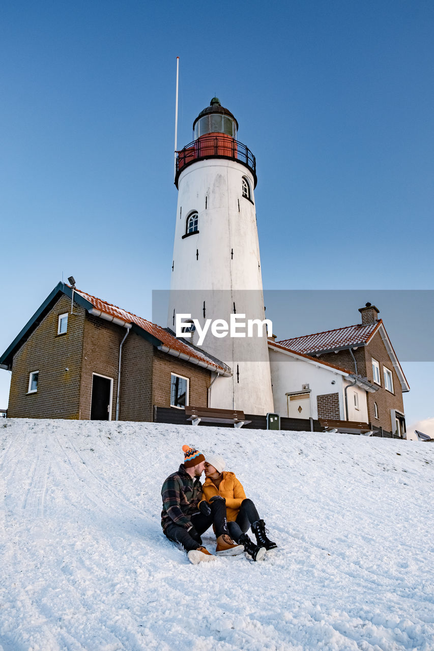 Full length of couple sitting on snow covered land against lighthouse