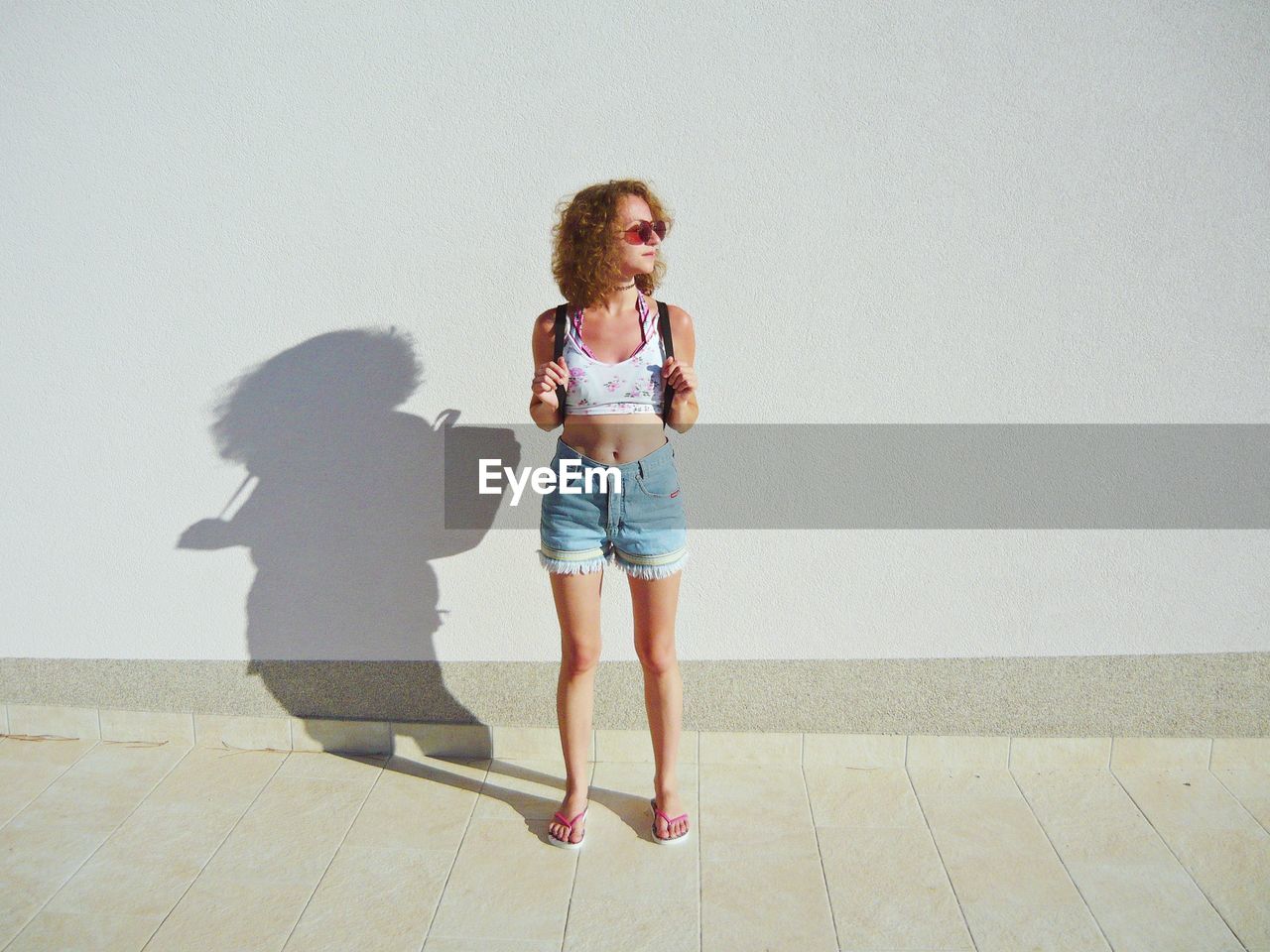 Full length of woman standing on sidewalk against white wall during sunny day