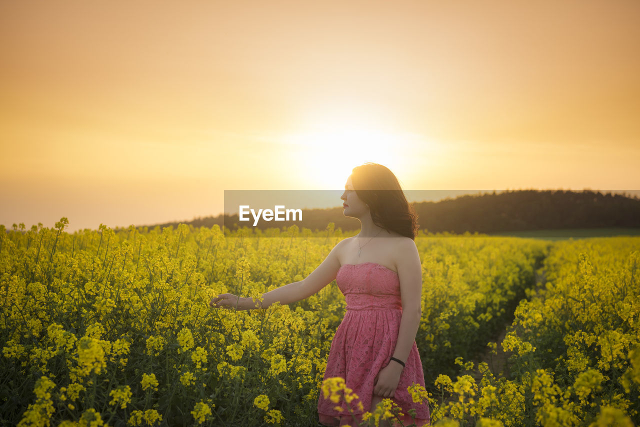 Woman standing amidst yellow flowering plants on field against sky during sunset