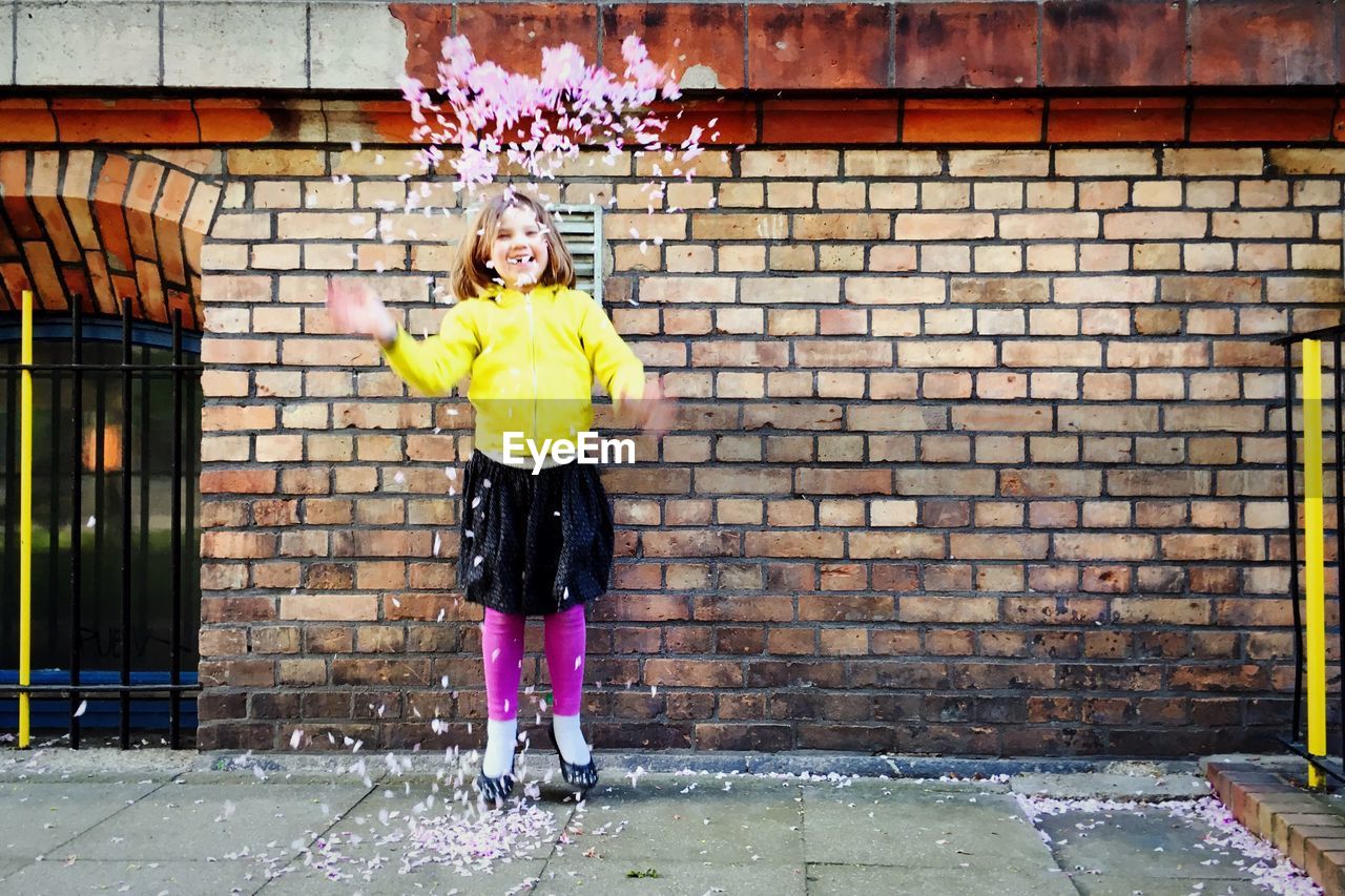 Portrait of cheerful girl throwing flower petals against brick wall