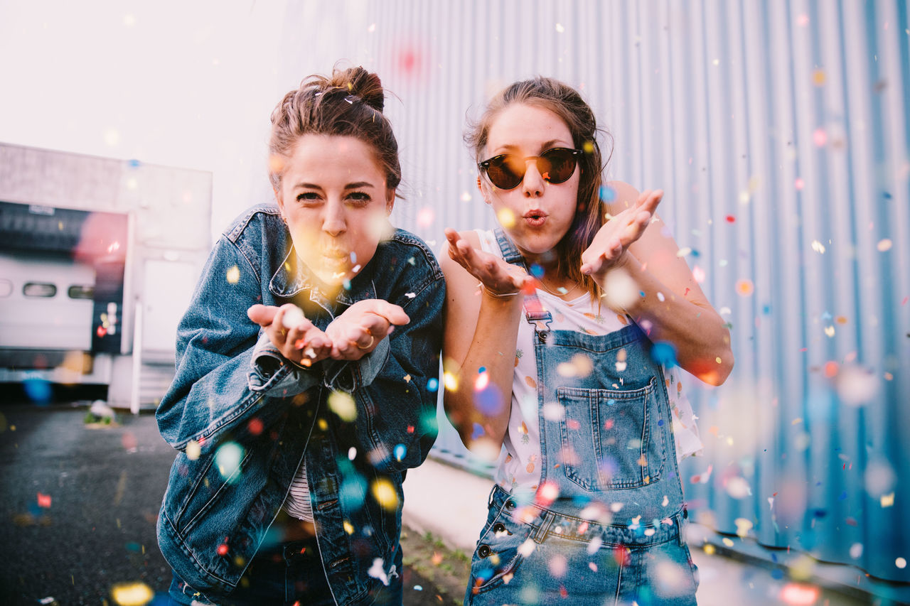 Portrait of young women blowing confetti while standing outdoors