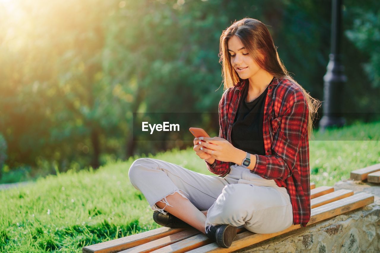 Smiling woman using mobile phone while sitting at bench in park