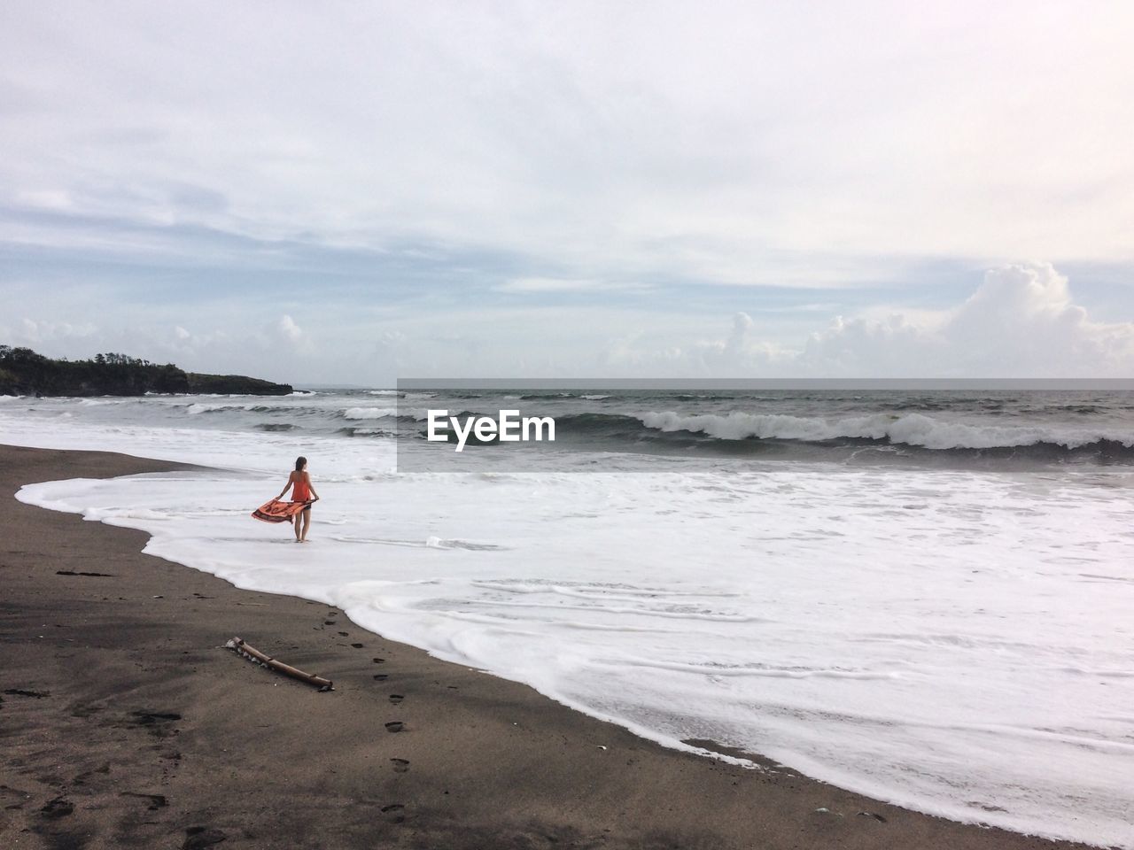 Woman holding scarf while standing at shore against cloudy sky