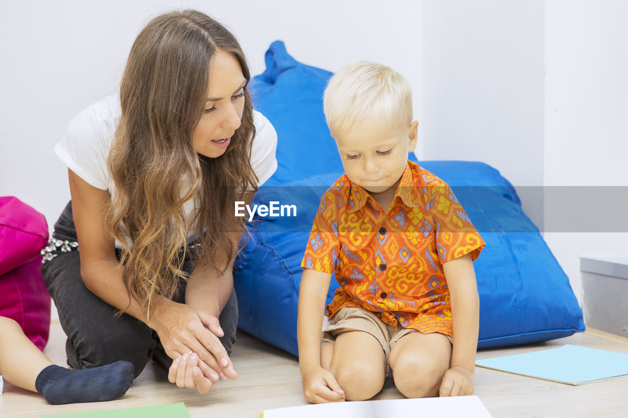 Mid adult teacher teaching student while sitting on floor against bean bag at school