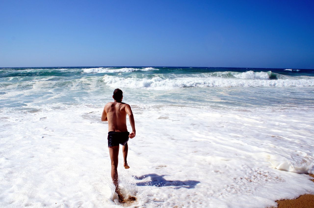 Rear view of shirtless man running on sea against clear blue sky