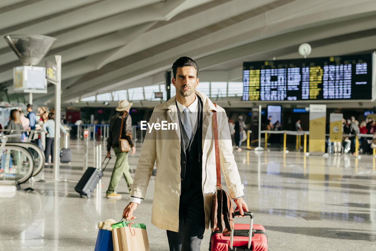 Crop of man with dark hair in stylish clothes walking with suitcase in terminal of airport