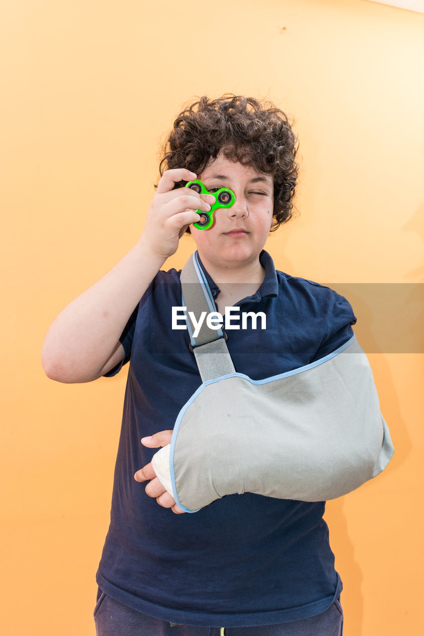 Boy with broken hand holding fidget spinner while standing against orange background