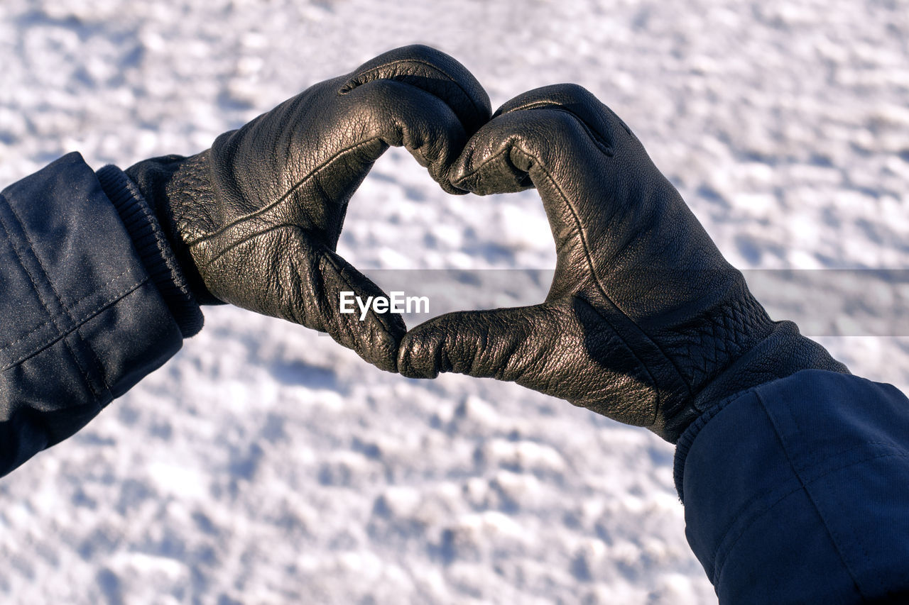 Male hands in black gloves are folded in shape of heart against background of white snow