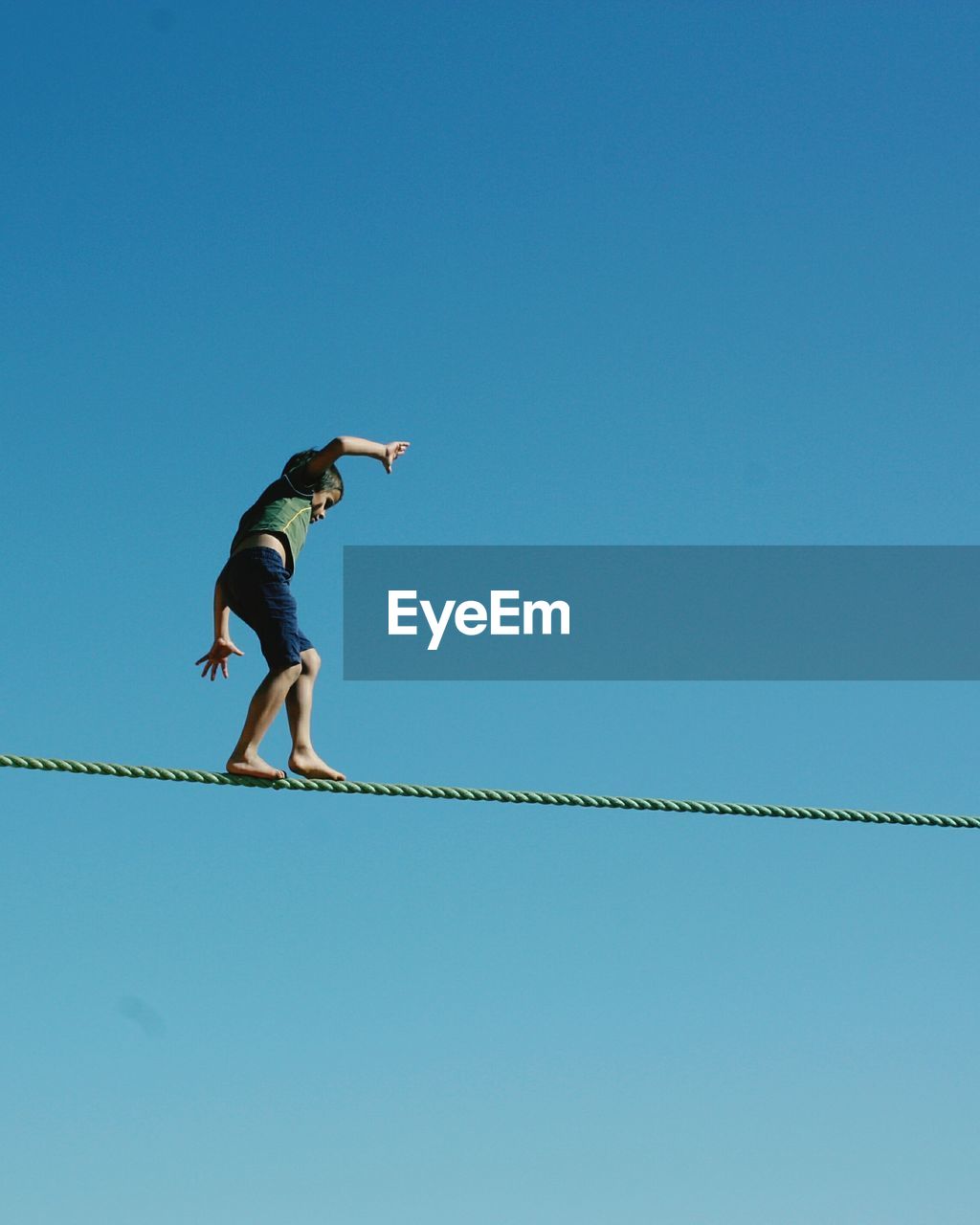 Low angle view of boy walking on rope against clear blue sky during sunny day