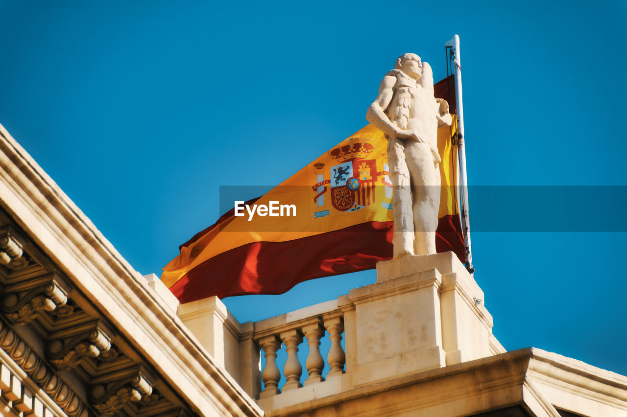 Low angle view of historical building and spanish national flag against blue sky, mnac, barcelona 