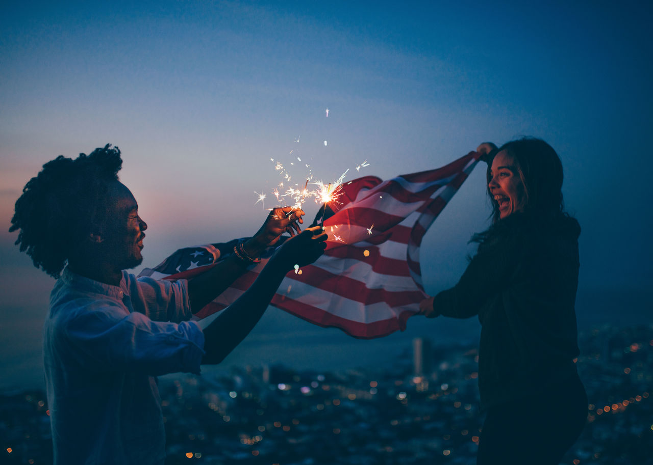 Side view of mid adult man burning sparklers while girlfriend holding american flag against sky at night