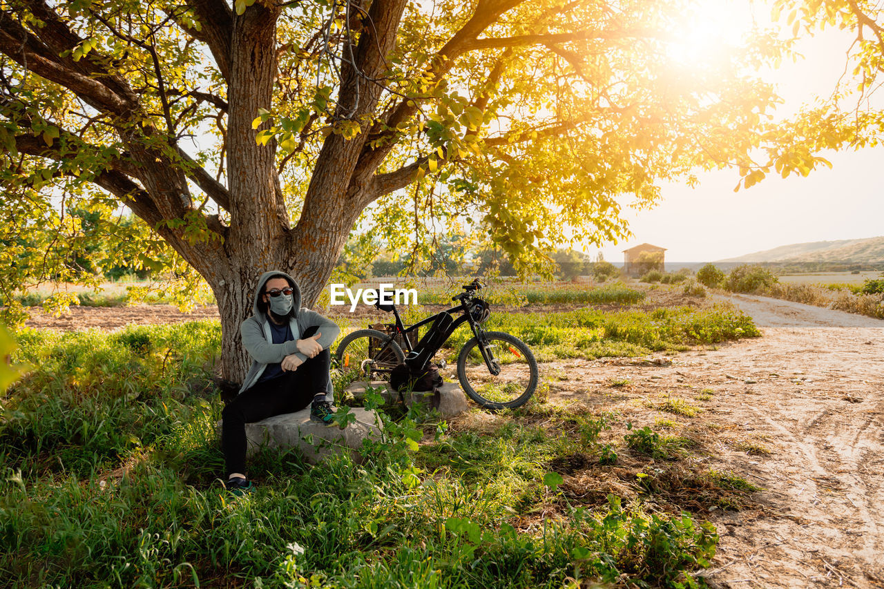 Man with bicycle sitting by tree on field against sky
